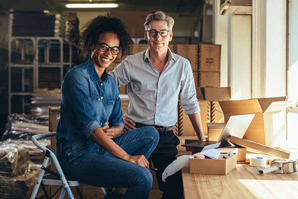 Smiling diverse business owners in an office setting, discussing finances and business growth, with packaging materials and a laptop on the table, representing small business collaboration and management.
