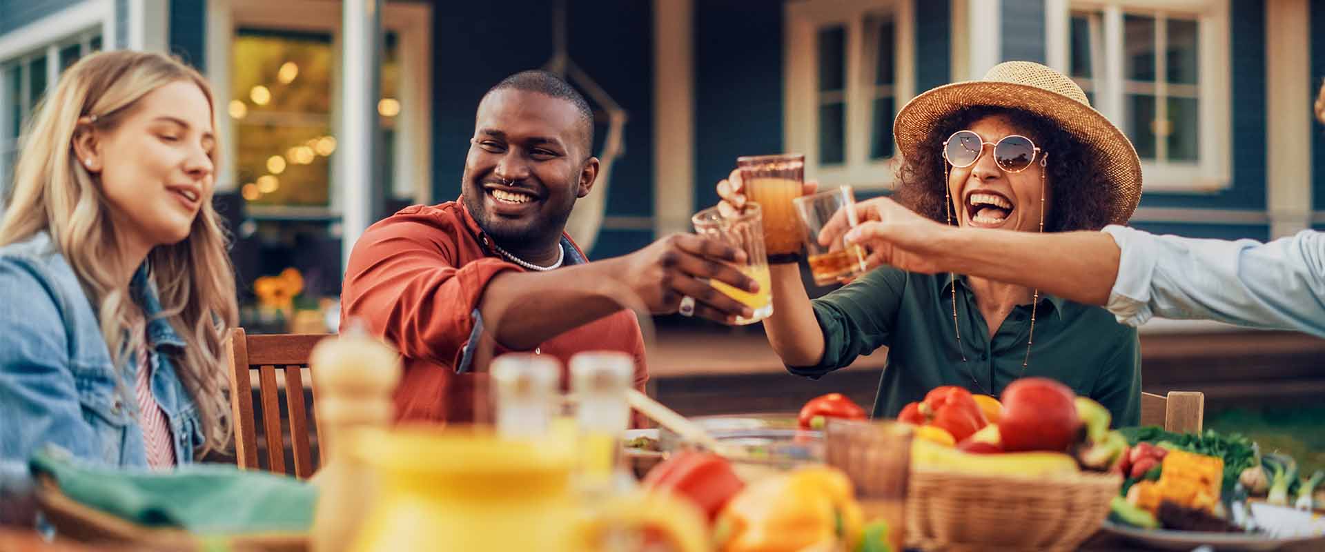Group of friends celebrating with drinks at a table filled with fresh fruits and food, enjoying a lively outdoor gathering.
