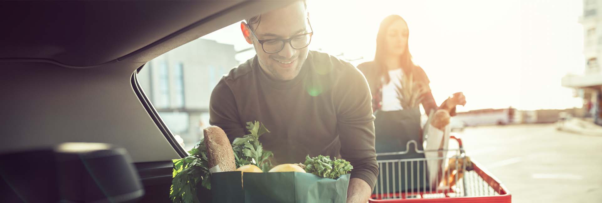 Man unloading groceries from a car, with a woman pushing a shopping cart in the background, highlighting everyday shopping experiences.