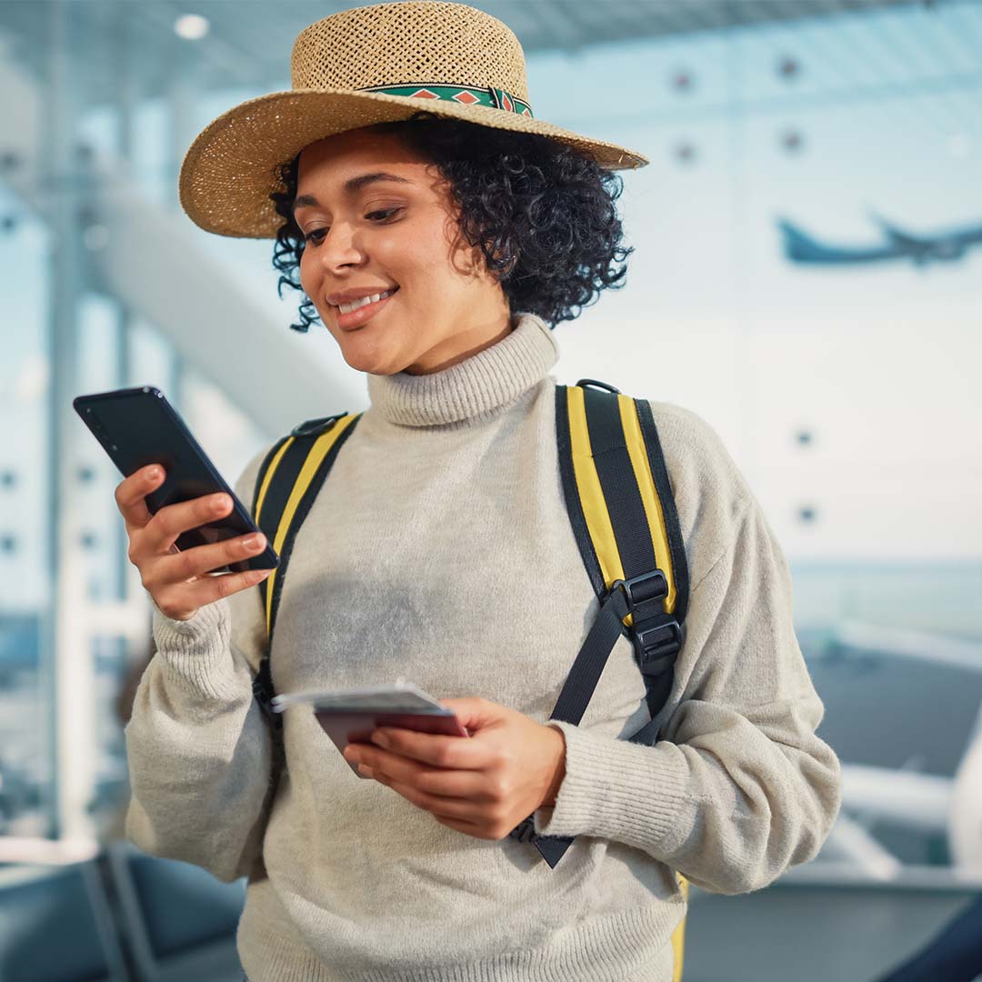 Woman in airport holding smartphone and notebook, preparing for travel, emphasizing the importance of submitting travel notices through M&F Bank's mobile app.