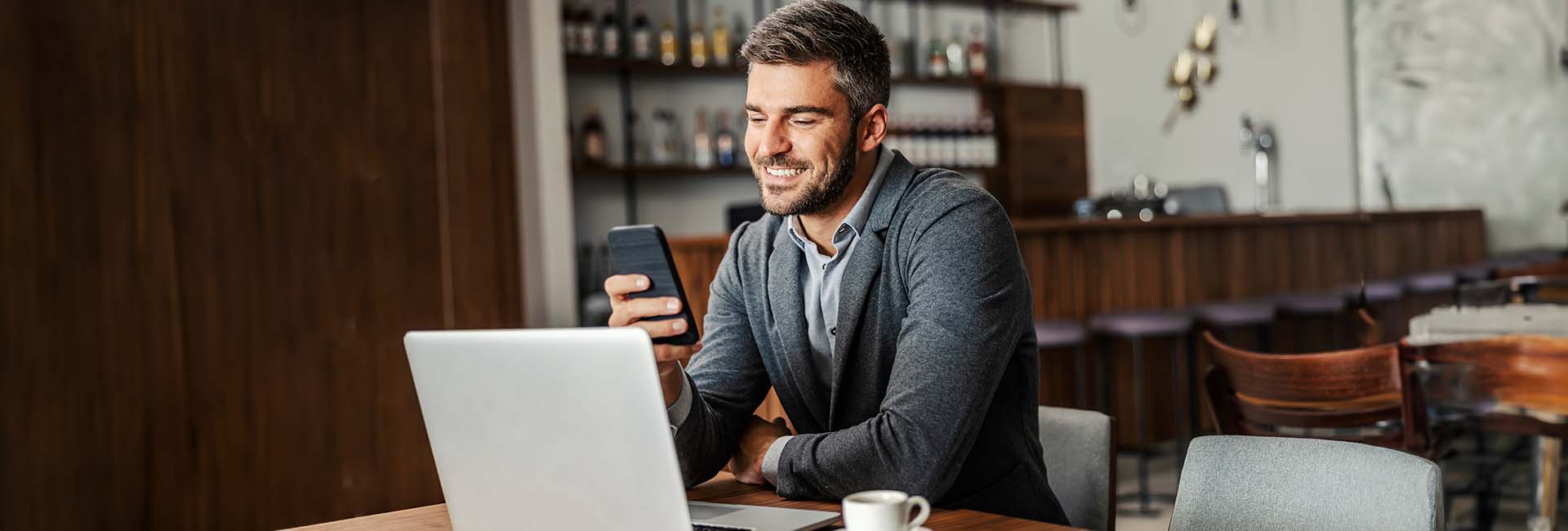 Man using smartphone and laptop in a caf&eacute;, illustrating seamless money transfer experience with Zelle&reg; through M&F Bank.