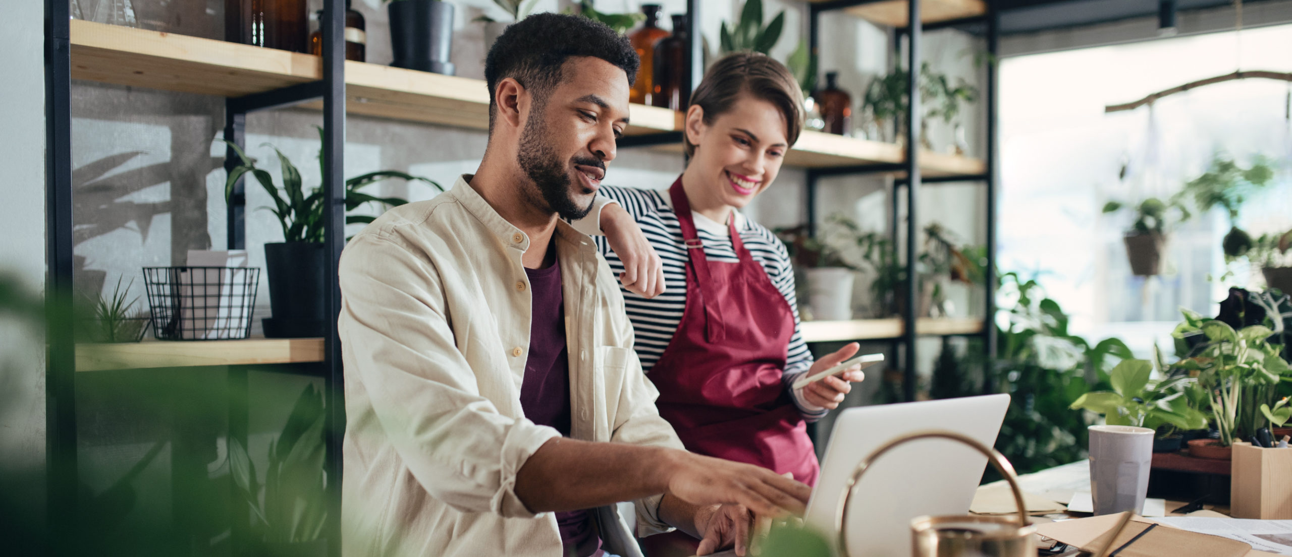 Two small business owners collaborating in a plant-filled workspace, one man working on a laptop and a woman in an apron smiling and holding a smartphone, illustrating teamwork and preparation for business success.