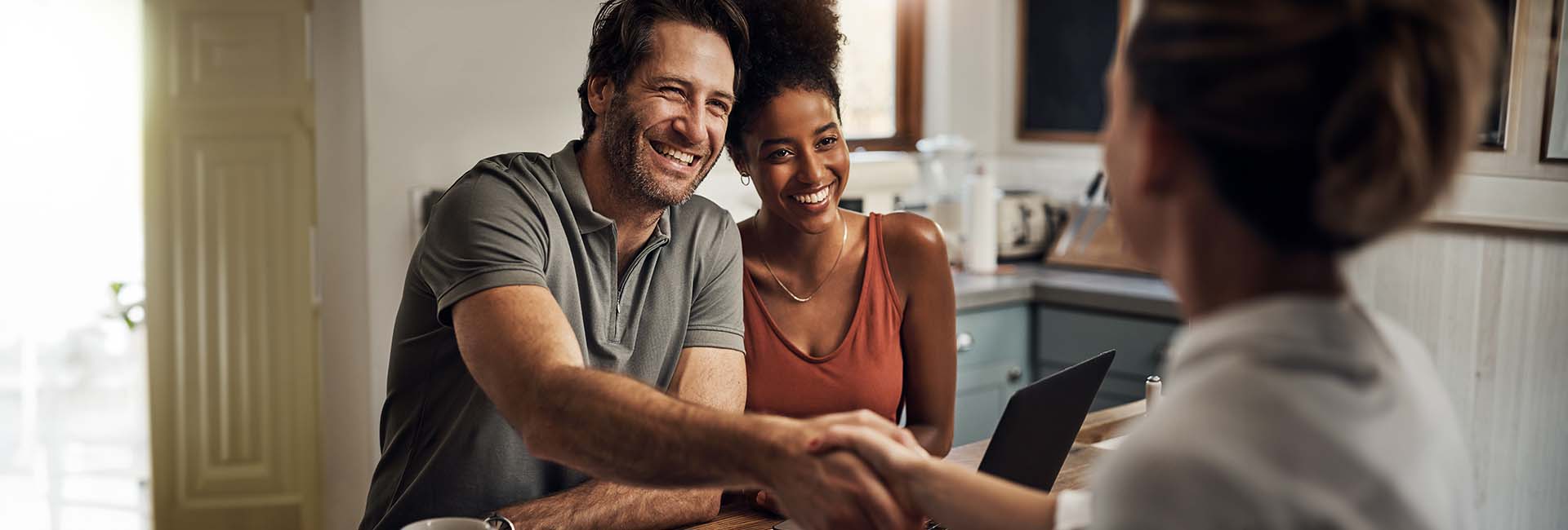 Couple smiling and shaking hands with a financial advisor in a cozy home setting, discussing investments and brokerage accounts.