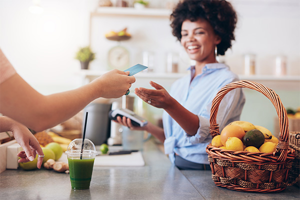 Customer using a credit card to pay at a caf&eacute; counter, with a smiling cashier and a basket of fresh fruits in the background, illustrating Clover Merchant Services in a business setting.