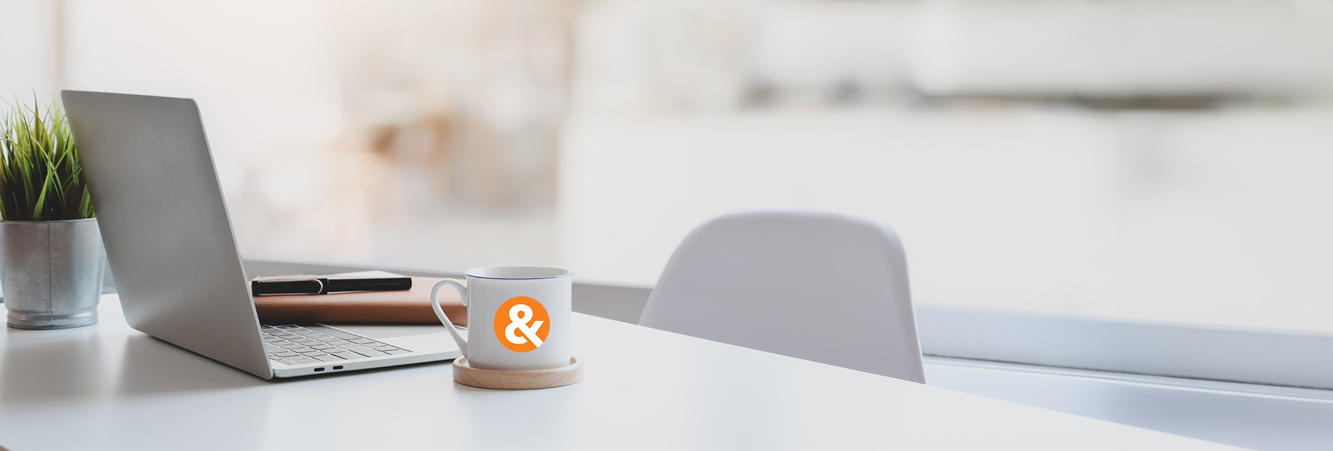 Laptop on a desk with a coffee mug featuring an ampersand symbol, representing M&F Bank's supportive work environment and team collaboration.